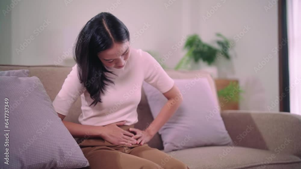 Concerned asian woman suffering from stomach-ache abdominal pain and colon problem or menstruation feels very painful with arms sitting on sofa the living room at her home. Concerned asian woman suffering from stomach-ache abdominal pain and colon problem or menstruation feels very painful with arms sitting on sofa the living room at her home.