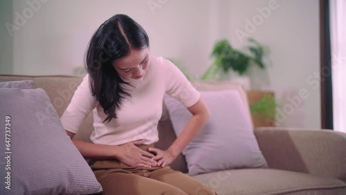 Concerned asian woman suffering from stomach-ache abdominal pain and colon problem or menstruation feels very painful with arms sitting on sofa the living room at her home. photo