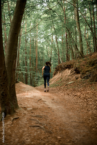 Rear view of girl with brown hair in pony tail running race trail competition on hilly forest area between tall deciduous trees © fesenko