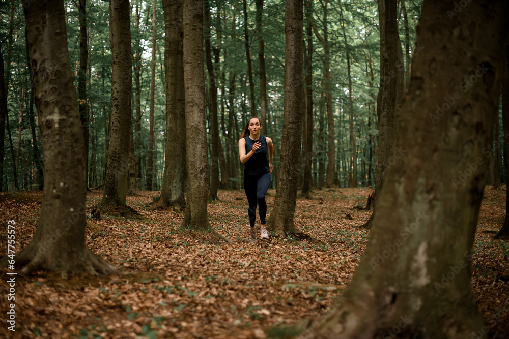 Young female athlete in black sportswear doing morning training in forest among trees.