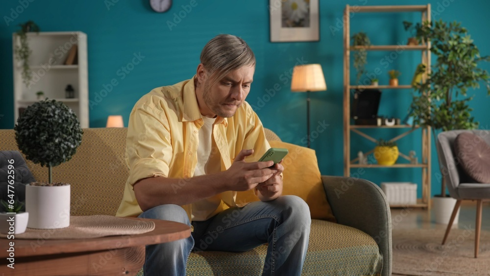 A man using a smartphone while sitting on the couch at home close up. A man scrolls through his feed on social networks, looks at photos, videos, and plays an online game.