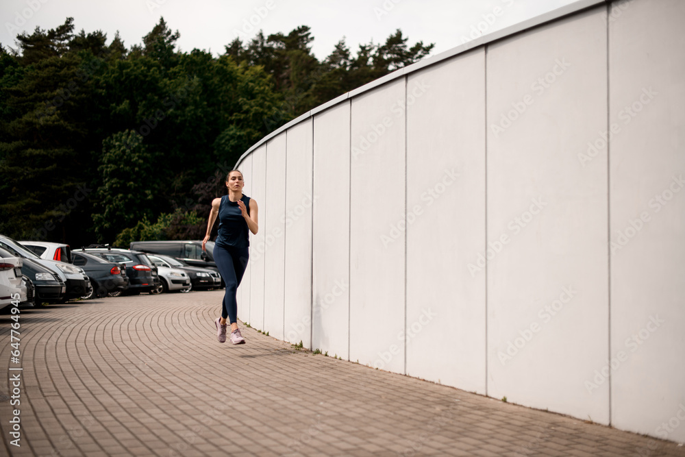 Young woman athlete running on foothpath in city street in front of white wall in morning