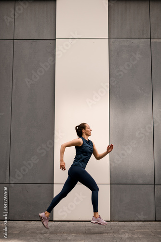 Beatutiful female athlete in sportswear jogging in city street against wall background