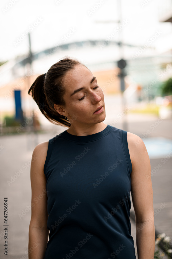 Fit healthy slim caucasian brunette in sportswear and with ponytail standing on city street with eyes closed