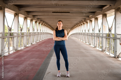 Outdoor shot of sporty woman dressed in activewear with thoughtful face expression and arms crossed on bridge