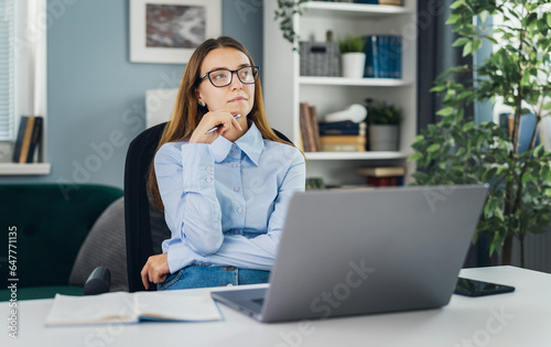 Young thoughtful woman wearing eyeglasses dreaming at desk with laptop, motivated idea concept, working in cozy light room