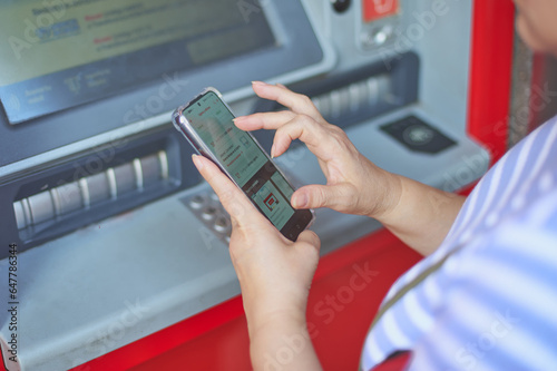 Fotografía Unrecognizable woman holding a cell phone in front of a bank ATM carrying out an