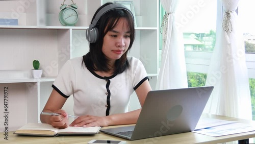 Beautiful young asian woman watching live video or video call of teacher teaching on laptop in her home, Take notes of important conversations and messages during the teacher's teaching.