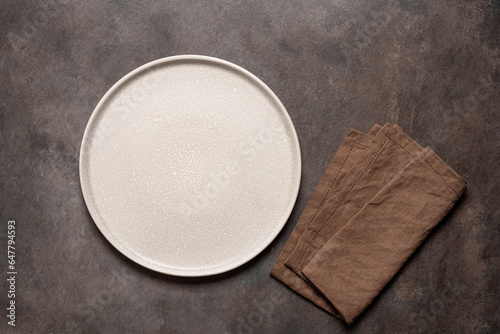 Empty beige plate with brown linen napkin on brown rustic background. Top view, flat lay.