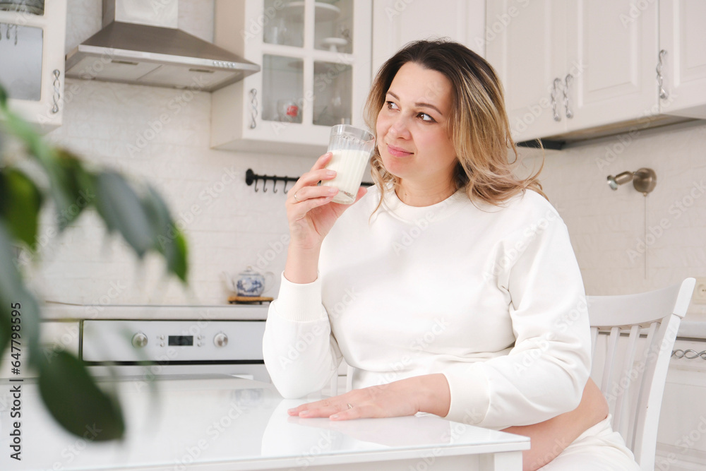 Beautiful pregnant woman in white suit is drinking milk in light kitchen. Beautiful pregnant woman in white suit is drinking milk in light kitchen.