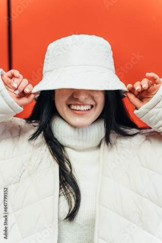 Happy funny carefree joyful young woman or teen girl with toothy smile puts on white cozy warm fluffy fuzzy artificial teddy lamb wool bucket hat that covers her eyes. Fashion concept Studio head