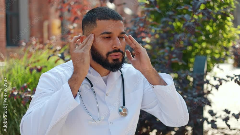 Arabian male doctor wearing white lab coat and stethoscope suffering from strong migraine and breathing fresh air outdoors. Sick man massaging temples in order to relief symptoms of headache. Arabian male doctor wearing white lab coat and stethoscope suffering from strong migraine and breathing fresh air outdoors. Sick man massaging temples in order to relief symptoms of headache.