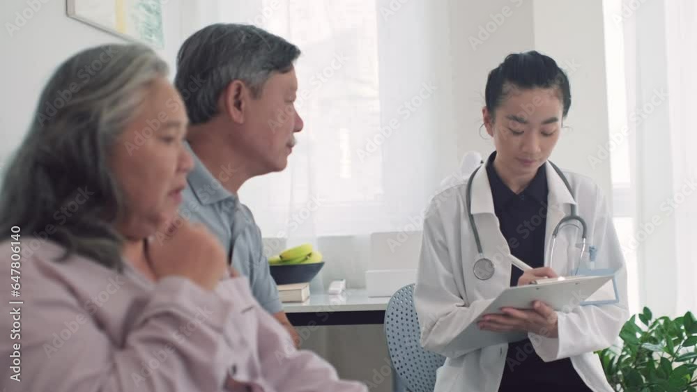Rack focus of young Asian doctor filing medical card of senior woman having asthma attack at home Rack focus of young Asian doctor filing medical card of senior woman having asthma attack at home