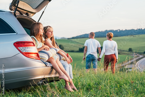 Two sisters girls sitting in car trunk enjoying sunset light. Young caucasian couple have auto trip break with hand in hand walk by green grass meadow. Family values, traveling and friendship concept.