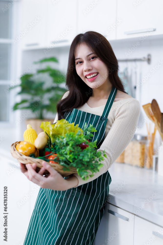 Happy portrait of young asian woman holding a basket of vegetables of standing a cheerful preparing food and enjoy cook cooking with vegetables, while standing on a kitchen Condo life or home Happy portrait of young asian woman holding a basket of vegetables of standing a cheerful preparing food and enjoy cook cooking with vegetables, while standing on a kitchen Condo life or home