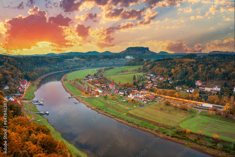 A beautiful autumn landscape of Elbe Valley in Saxon Switzerland, Germany. The river, hills, and a small town Rathen at the dawn with a dramatic sunrise in the background.