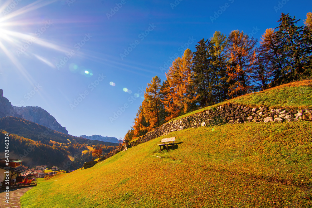 A beautiful autumn landscape of the Dolomite Alps in Val Gardena, Italy. Hillside with yellowed larches, stone wall and wooden bench A beautiful autumn landscape of the Dolomite Alps in Val Gardena, Italy. Hillside with yellowed larches, stone wall and wooden bench