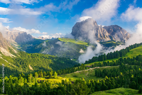 A vibrant depiction of the Sassolungo Massif and Gardena Valley in Dolomite Alps, Italy. The foreground showcases a lush green valley, while the background highlights a cloud-covered mountain peaks