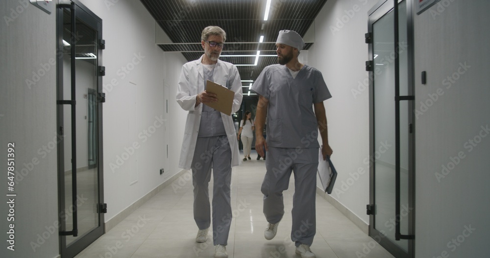 Mature doctor with colleague walk together the clinic corridor. Healthcare professionals discuss treatment, diagnosis or medical test results. Medical staff and patients in hospital or medical center.
