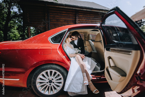 Stylish groom and beautiful bride in sunglasses hugging while sitting in a red car. Wedding photography, portrait, lifestyle.