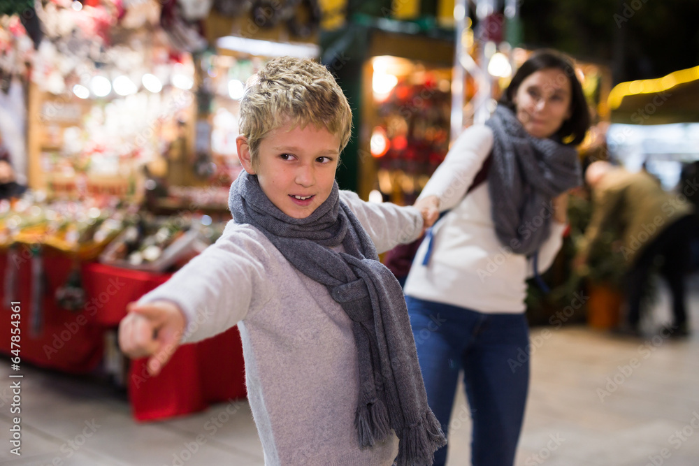 Preteen boy pulling his mom hand, demanding to buy something for him at outdoor Christmas market