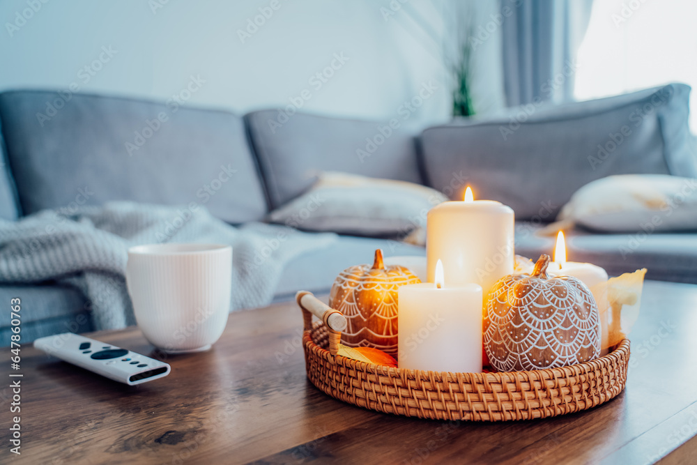 Autumn fall cozy mood composition for hygge home decor. Orange pumpkins, burning candles, cup with hot drink and remote controller on coffee table in living room. Movie night at home. Cozy relax time.