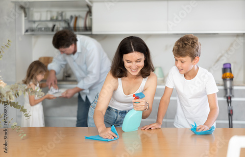 Positive mother and son cleaning table surface, father and son doing dishwashing in the kitchen