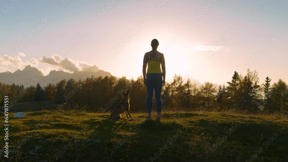 CLOSE UP, LENS FLARE: Lady with a dog admires spectacular sunset at the hilltop. She is rewarding herself with an outstanding view of high mountains in golden light after trail running up the hill. CLOSE UP, LENS FLARE: Lady with a dog admires spectacular sunset at the hilltop. She is rewarding herself with an outstanding view of high mountains in golden light after trail running up the hill.