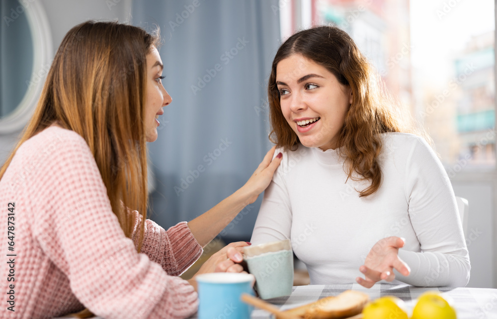 Smiling young woman with cup of tea talking with girlfriend at home