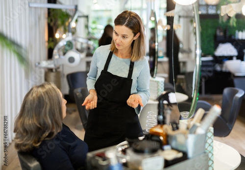 Portrait of young woman professional hair stylist talking to elderly female client in salon, choosing new hairdo .