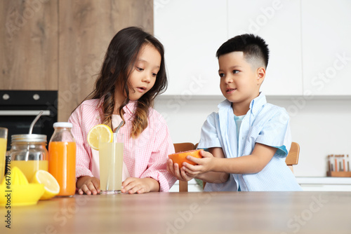 Little Asian children with glass of fresh citrus juice and grapefruit slice sitting at table in kitchen