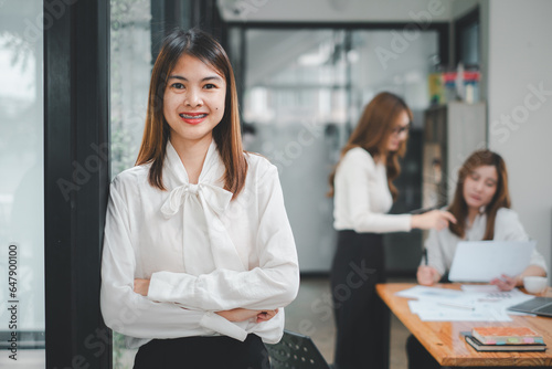 Happy business woman standing competently and smiling in open plan office with blurry collea gues sitting in office as background, Business concept. photo