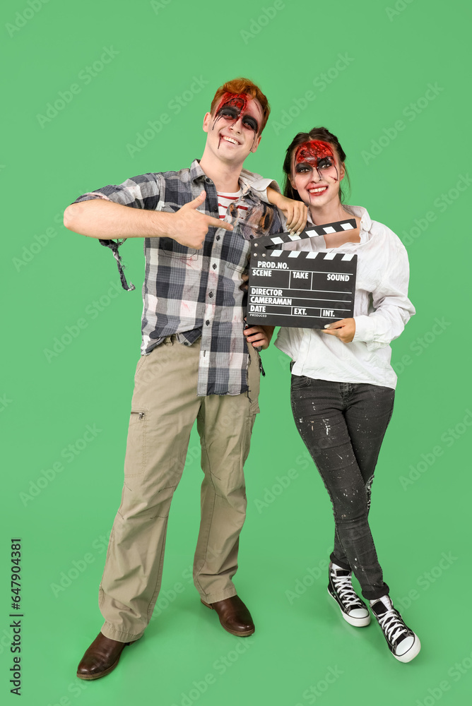 Fototapeta premium Young couple dressed for Halloween as zombies with movie clapper on green background