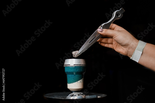 The hand of a girl with a special tool with tongs puts coal in a ceramic hookah bowl, an object to smoke in the dark background
