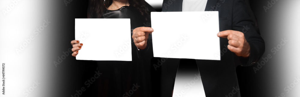 Two people, a man and a woman, holding a sheet of A4 paper in their hands, an empty space for text and design white blank on a dark background Two people, a man and a woman, holding a sheet of A4 paper in their hands, an empty space for text and design white blank on a dark background