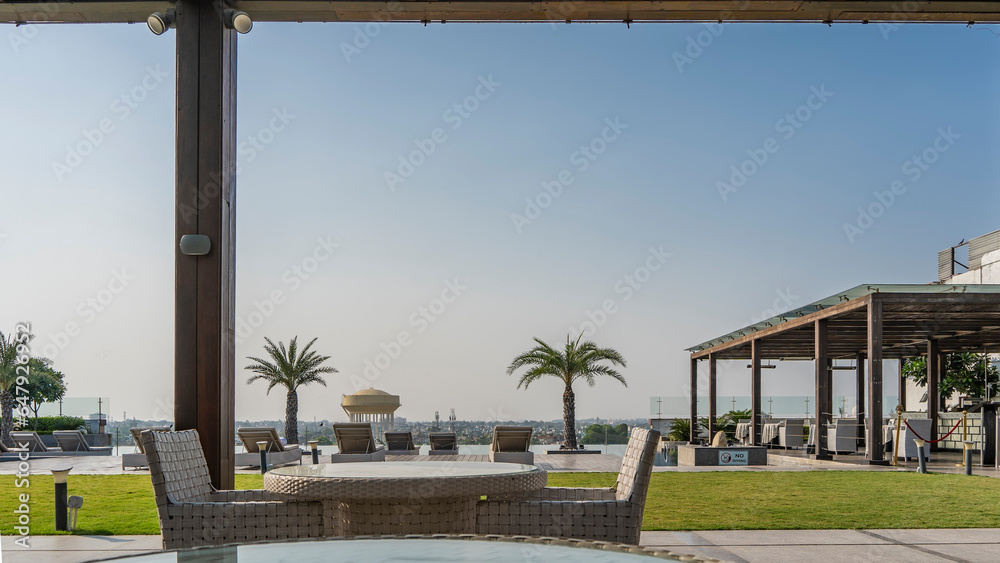 A relaxation area on the roof. Sun beds are located along the infinity pool. Palm trees against the blue sky. The city is visible in the distance. In the foreground - a round table and wicker chairs A relaxation area on the roof. Sun beds are located along the infinity pool. Palm trees against the blue sky. The city is visible in the distance. In the foreground - a round table and wicker chairs