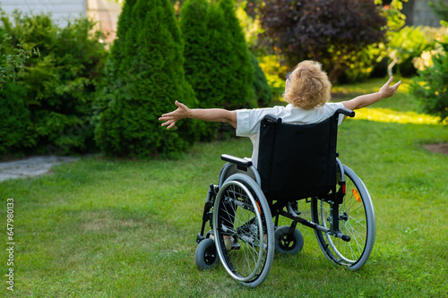 Rear view of an elderly woman spread her arms to the sides while sitting in a wheelchair on a walk outdoors. 