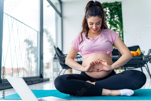 Free Download young pregnant woman siting on a yoga mat making a heart shape on her pregnant belly, excited young mother concept - GFXLoad