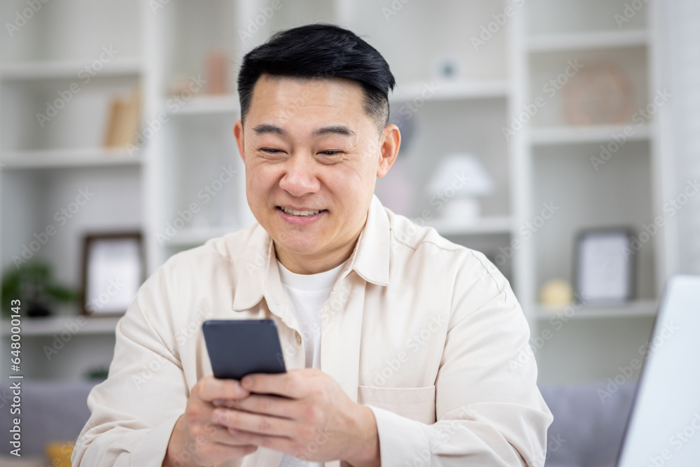 Mature Asian man sitting at table at home, man using phone, typing message on smartphone, app user and browsing internet in home cinema room, smiling contentedly. Mature Asian man sitting at table at home, man using phone, typing message on smartphone, app user and browsing internet in home cinema room, smiling contentedly.