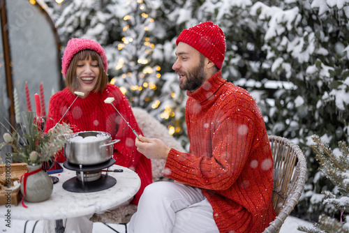 Man and woman have romantic dinner with fondue, while sitting together by the table at beautifully decorated snowy backyard. Young family celebrating winter holidays outdoors photo