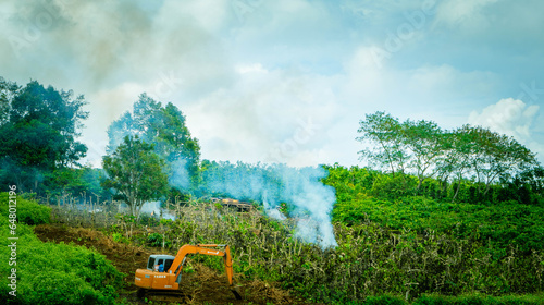 Yellow crawler excavator digs the ground against the blue sky with clouds, green trees and smoke. Construction work in the highlands.