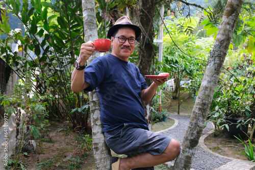 Asian guy leaning on a palm tree in the garden, drinking hot coffee in red ceramic cup photo