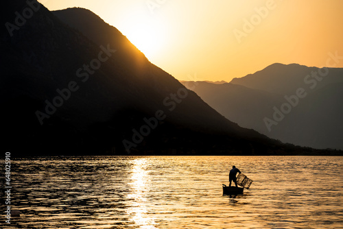 At sunset, amidst the mountains, a man is fishing in a small boat in the bay. Silhouettes in the golden hour. Dramatic scene.