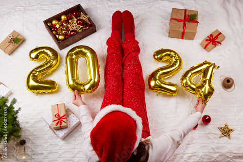 Happy New Year 2024. Golden balloons with numbers. The girl is sitting on the bed and holding helium balloons in her hands. Comfort in the house on Christmas Eve, gift wrapping.