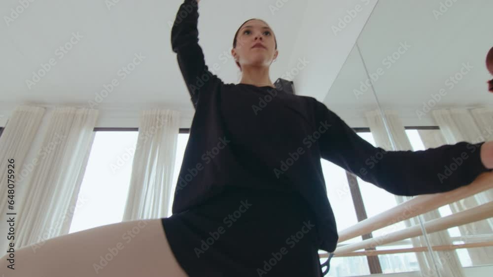 Young talented ballerina doing passe on demi pointe, taking one arm off the barre and keeping a balance while practicing in a dance studio. Tilt-up shot, low angle