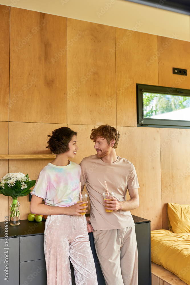 happy multicultural couple standing in pajamas and holding glasses with orange juice in morning happy multicultural couple standing in pajamas and holding glasses with orange juice in morning