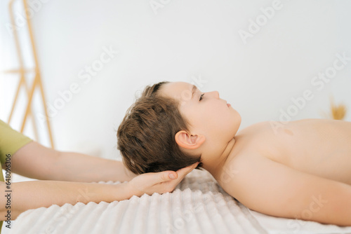 Close-up side view of unrecognizable professional female osteopath doing neck adjustment to five year old boy patient in physiotherapist cabinet, during manual therapy. Concept of healthy childcare. photo