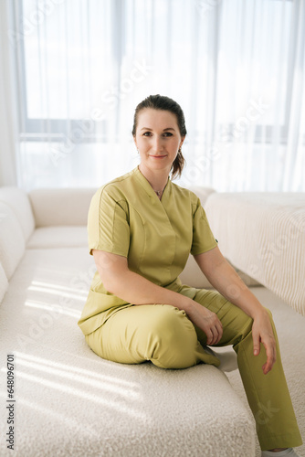 Vertical portrait of smiling female masseuse therapist in uniform sitting posing on couch looking at camera with friendly expression, on background of window in wellness center. Concept of body care.