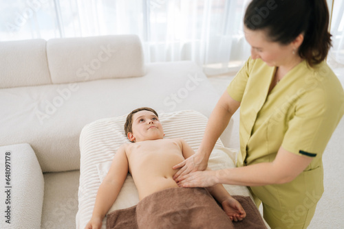 Top view of female pediatric masseuse making therapeutic massage on tummy to five year old boy in clinic. Rehabilitation massage on stomach of child lying on massage table. Concept of childcare.