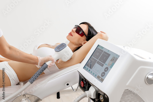 Body care concept. Laser hair removal. Young Caucasian woman in safety glasses lies on the sofa, female cosmetologist doing laser hair removal in a beauty salon